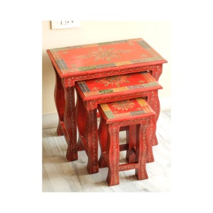 May include: A set of three nesting tables with intricate hand-painted designs. The tables are made of wood and have a red finish. The designs feature floral motifs and geometric patterns in shades of gold, green, and blue.