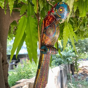 May include: A colorful wooden bird ornament with intricate details and a hanging loop. The bird is painted in shades of blue, green, yellow, red, and orange, with a paisley pattern on its body.
