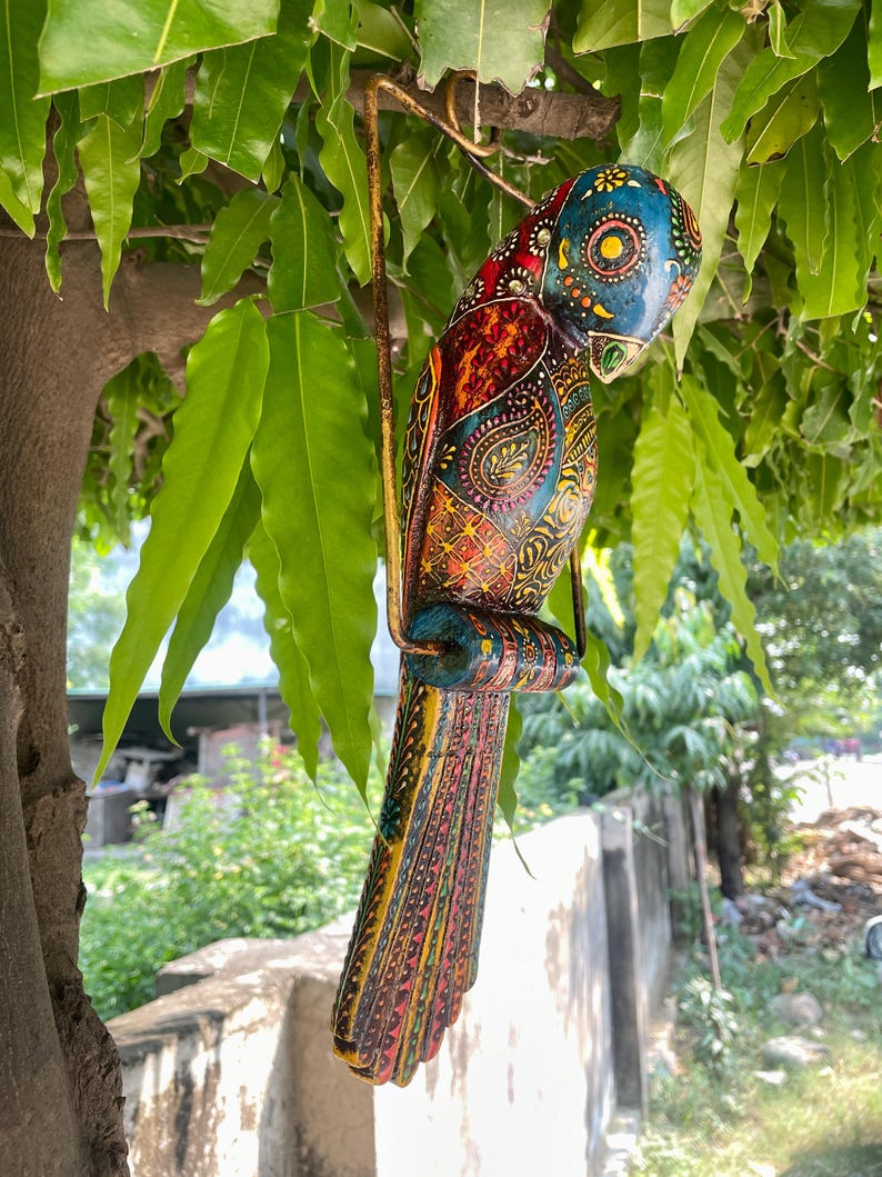 May include: A colorful wooden bird ornament with intricate details and a hanging loop. The bird is painted in shades of blue, green, yellow, red, and orange, with a paisley pattern on its body.