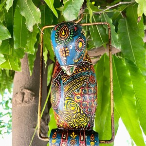 May include: A colorful, hand-painted wooden bird ornament with intricate details. The bird is blue, yellow, red, green, and black with a gold metal ring.