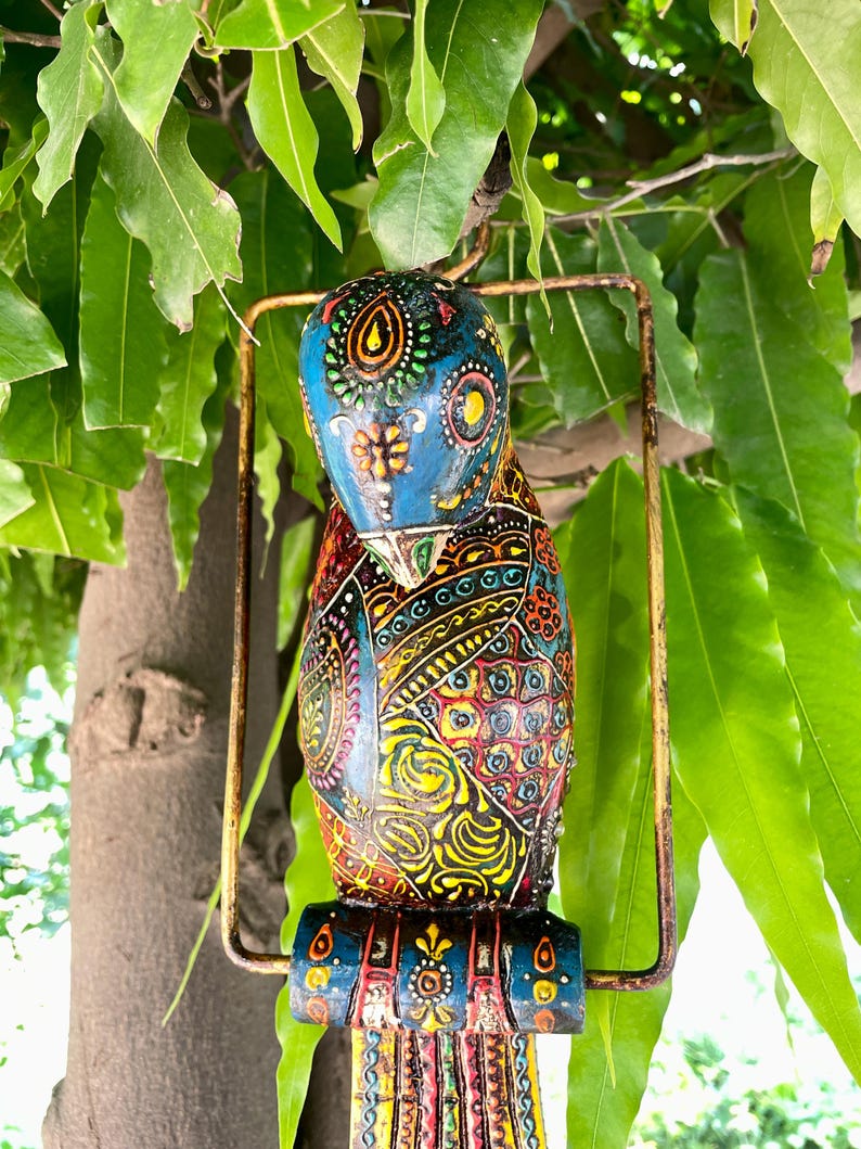 May include: A colorful, hand-painted wooden bird ornament with intricate details. The bird is blue, yellow, red, green, and black with a gold metal ring.