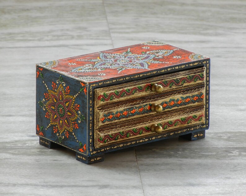 Puede incluir: Una peque&ntilde;a caja de madera con tres cajones, pintada en azul, naranja y dorado con intrincados dise&ntilde;os florales. La caja tiene pomos de lat&oacute;n en los cajones.