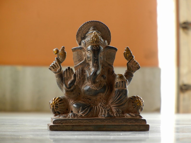 Puede incluir: Una estatua de color marr&oacute;n oscuro del dios hind&uacute; Ganesha, sentado con la trompa levantada y sosteniendo un objeto peque&ntilde;o en la mano. La estatua est&aacute; decorada con detalles dorados y se encuentra sobre una base de madera.