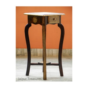 May include: A small, dark wood side table with a single drawer and ornate gold accents. The table has three legs with a curved design.