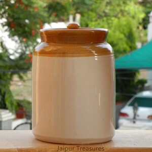 May include: A white ceramic jar with a brown lid. The jar is sitting on a wooden surface. The text "Jaipur Treasures" is printed on the surface.