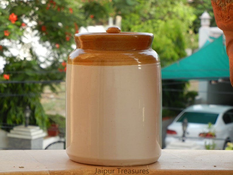 May include: A white ceramic jar with a brown lid. The jar is sitting on a wooden surface. The text "Jaipur Treasures" is printed on the surface.