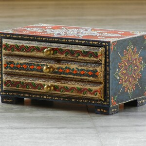 Puede incluir: Una peque&ntilde;a caja de madera con tres cajones pintados en un dise&ntilde;o azul, rojo, naranja y dorado. La caja tiene un patr&oacute;n floral en la parte delantera y detalles dorados en los cajones.