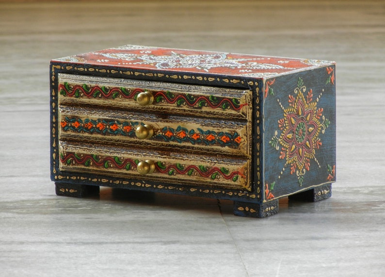 Puede incluir: Una peque&ntilde;a caja de madera con tres cajones pintados en un dise&ntilde;o azul, rojo, naranja y dorado. La caja tiene un patr&oacute;n floral en la parte delantera y detalles dorados en los cajones.
