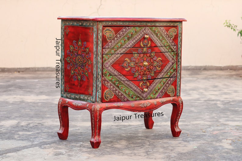 May include: A red wooden chest of drawers with three drawers. The drawers are decorated with intricate floral patterns in shades of green, gold, and blue. The chest is accented with silver trim and has a distressed finish. The chest is labeled "Jaipur Treasures" on the front and side.