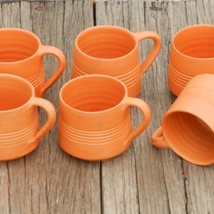 May include: Six terracotta mugs with handles. The mugs are a light orange color and have a textured, ribbed surface.