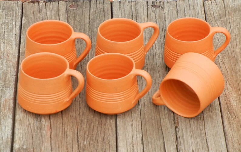 May include: Six terracotta mugs with handles. The mugs are a light orange color and have a textured, ribbed surface.