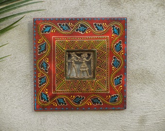 Könnte beinhalten: Ein roter Holzrahmen mit einem dekorativen Rand aus blauen, grünen und gelben Blättern. Der Rahmen enthält ein Relief aus Messing mit zwei tanzenden Figuren.