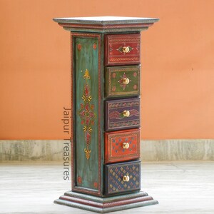 K&ouml;nnte beinhalten: Ein blaugr&uuml;ner Holzschrank mit sechs Schubladen. Jede Schublade ist mit einem anderen farbenfrohen Design bemalt und hat einen Messingknopf. Der Schrank steht auf einem hellbraunen Boden. Der Text "Jaipur Treasures" ist an der Seite des Schranks aufgedruckt.