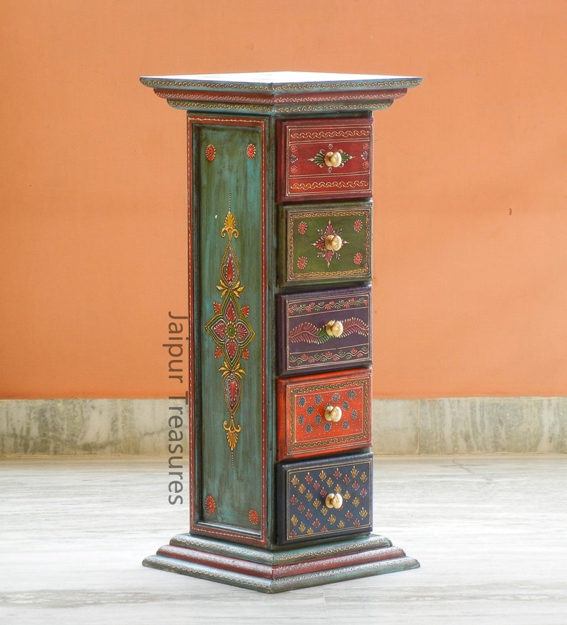 K&ouml;nnte beinhalten: Ein blaugr&uuml;ner Holzschrank mit sechs Schubladen. Jede Schublade ist mit einem anderen farbenfrohen Design bemalt und hat einen Messingknopf. Der Schrank steht auf einem hellbraunen Boden. Der Text "Jaipur Treasures" ist an der Seite des Schranks aufgedruckt.