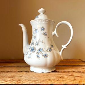 Peut inclure: Théière en céramique blanche avec couvercle, ornée d'un motif floral de fleurs bleues et de feuilles grises. La théière a un bec verseur incurvé et une grande anse arquée. Le design est de style classique et vintage.