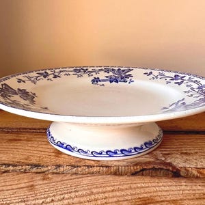 Op de afbeelding: Witte keramische taartstandaard met blauw bloemmotief. De ovale plaat staat op een voetstuk. De rand en de basis zijn versierd met blauwe bloempatronen. De standaard staat op een houten oppervlak.