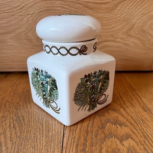 Peut inclure: Pot en céramique blanche avec un couvercle, orné d'un motif floral d'herbes vertes, bleues et brunes liées par un ruban doré. Le pot a un motif de corde dorée autour du haut.