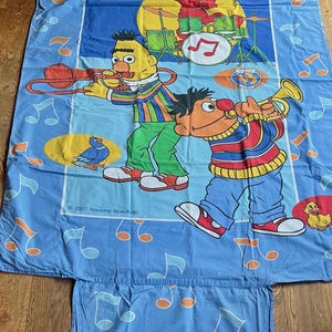 Op de afbeelding: Een lichtblauw laken met een Sesame Street muziekthema met Elmo op drums, Bert op trombone en Ernie op trompet. Het ontwerp bevat muzieknoten, een duif, een goudvis en een badeendje. De tekst op het laken luidt: 2001 Sesame Workshop.