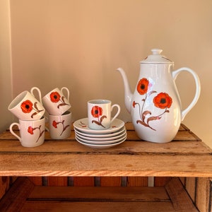 Op de afbeelding: Een witte keramische koffiepot met een bloemenontwerp met oranje klaprozen. De set bevat vier bijpassende kopjes en schoteltjes.
