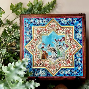 Op de afbeelding: Vierkante, omlijste keramische tegel met een gedetailleerd vogel- en bloemmotief. De tegel heeft een centraal stervormig beeld van een vogel bij water, omgeven door een blauwe en bloemenrand. Het frame is gemaakt van donker hout.
