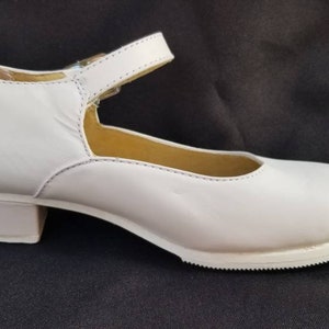 May include: White leather shoe with a small heel and an adjustable strap. The shoe has a rounded toe and a textured sole. The shoe is designed for dance or formal wear.