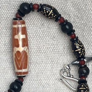 May include: A handcrafted necklace featuring a central cylindrical bead with a carved design in cream and brown. The necklace incorporates black and red beads, with gold-coloured accents on some of the black beads. A silver-toned clasp secures the necklace.