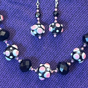 May include: A black beaded necklace and matching earrings set. The necklace and earrings feature large, round black beads with pink and white polka dots, interspersed with smaller black and pink beads. The jewelry is displayed on a purple textured surface.