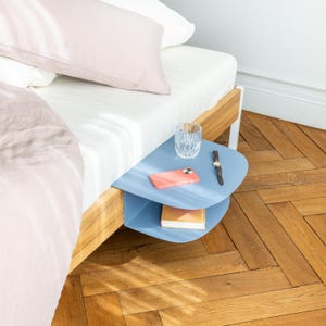 Pode incluir: Uma mesa de cabeceira azul clara presa a uma estrutura de cama de madeira. Um copo transparente, um telefone cor-de-coral, um relógio e um livro estão sobre a mesa. A cama tem roupa de cama branca e rosa. O chão é de madeira.