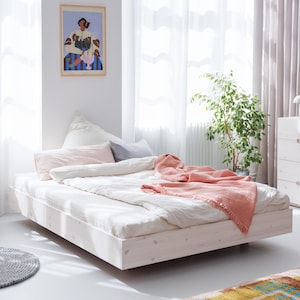 Op de afbeelding: Een modern, laag bedframe in een lichte houten afwerking. Het bed is opgemaakt met wit beddengoed, inclusief een dekbed en kussens. Een koraalkleurige sprei is over het voeteneinde van het bed gedrapeerd. Een ingelijst kunstwerk hangt aan de muur.