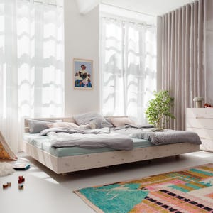 Op de afbeelding: Een lichtgekleurd houten bedframe met een lichtblauwe matras en grijs beddengoed. Het bed staat in een lichte kamer met witte gordijnen en een kleurrijk tapijt. Een kleine boom en een ingelijste foto zijn ook zichtbaar.