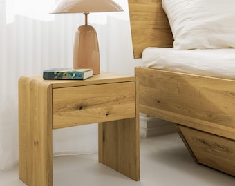 Könnte beinhalten: Ein helles Nachttischchen aus Holz mit einer Schublade und geschwungenem Design. Eine pfirsichfarbene Lampe mit rundem Schirm steht darauf, zusammen mit einem Buch. Ein hölzerner Bettrahmen ist teilweise im Hintergrund zu sehen.