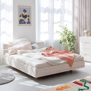 Op de afbeelding: Een licht houten bedframe met een matras en kussens, bedekt met een wit dekbed en een koraalkleurige sprei. Een ingelijst kunstwerk hangt aan de muur boven het bed. Een groene plant en een witte ladekast staan ook in de kamer.
