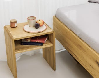 Könnte beinhalten: Ein helles Nachttischchen aus Holz mit einem unteren Regal, das Bücher enthält. Oben befinden sich ein kleines Glas, eine Tasse, ein Teller mit Obst und ein Bettrahmen. Der Tisch steht neben einem Bett mit weißer Bettwäsche.
