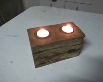 May include: A rustic wooden candle holder with two lit tea lights. The holder is made of stacked pieces of wood with a natural brown finish.