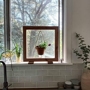 May include: A wooden framed window with a hanging flower pot. The pot is suspended by a rope and contains a small plant with red flowers. The window is set against a backdrop of trees and greenery.