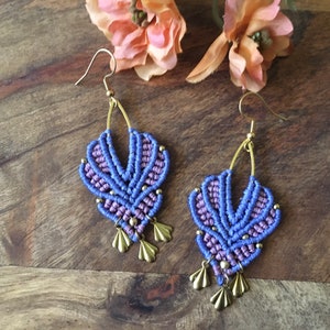 Puede incluir: Un par de pendientes de macramé morado y lavanda con detalles dorados y un borla. Los pendientes cuelgan de ganchos dorados.