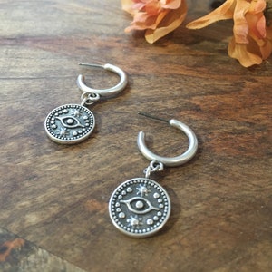 May include: A pair of silver hoop earrings with a charm hanging from each hoop. The charms are round and feature a silver eye with a black pupil and white details. The charms are surrounded by a circle of small silver dots.