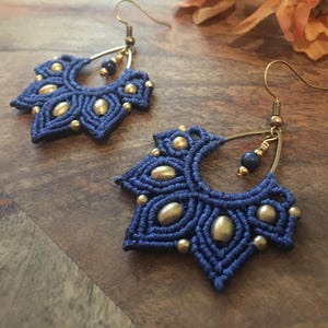 Könnte beinhalten: Ein Paar blaue und goldene Ohrringe mit einem blütenförmigen Design. Die Ohrringe sind aus Makramee gefertigt und haben goldene Perlen und einen Lapislazuli-Stein.