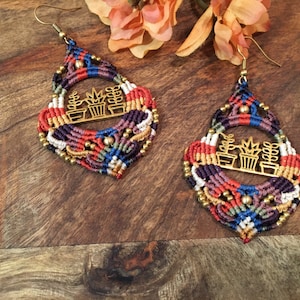 May include: A pair of colourful macrame earrings with a gold metal design of potted plants. The earrings are hanging from a wooden surface.