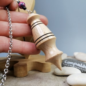 May include: A wooden pendulum with a pointed tip and two black bands around the middle, hanging from a silver chain.
