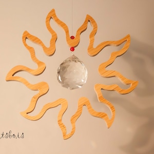 Peut inclure: Une décoration murale en forme de soleil en bois avec une boule de cristal suspendue au centre. Le soleil est en bois brun clair et a une texture légèrement ondulée. La boule de cristal est transparente et réfléchit la lumière.