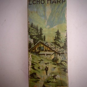 Könnte beinhalten: Vintage-Box für ein Musikinstrument The Echo Harp. Die Box zeigt eine farbenfrohe Illustration einer Bergszene mit einer kleinen Hütte, Bäumen und einer Person, die auf einem Weg geht. Der Text "The Echo Harp" ist in Schwarz auf der Box gedruckt, zusammen mit dem Namen des Herstellers "M. Hohner" und "Made in Germany".