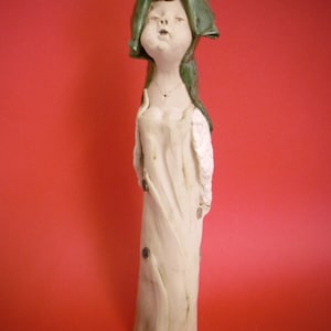 Könnte beinhalten: Eine Keramikfigur einer Person, die einen grünen Kapuzenumhang und ein weißes Kleid trägt. Die Figur steht auf einem roten Hintergrund.