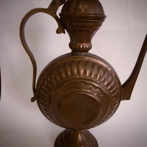 May include: A copper teapot with a round body and a decorative handle. The teapot has a lid with a finial and a spout. The teapot is on a stand with a round base.