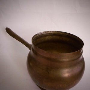 Op de afbeelding: Een vintage koperen pot met een lange handgreep. De pot heeft een rond lichaam en een licht uitlopende rand.