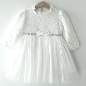 May include: White lace dress with a tulle skirt and a jeweled belt. The dress has long sleeves and a bow at the waist.