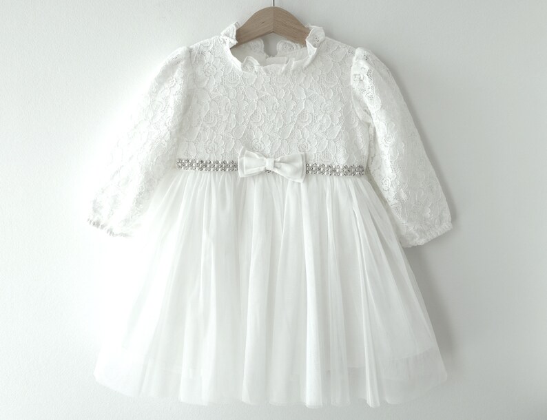 May include: White lace dress with a tulle skirt and a jeweled belt. The dress has long sleeves and a bow at the waist.