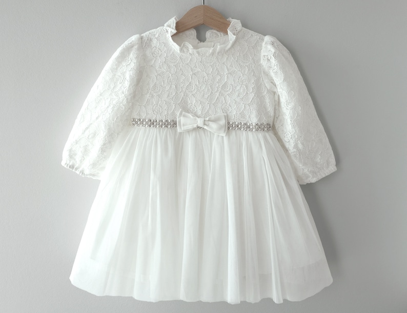 May include: White lace dress with a tulle skirt and a beaded belt with a bow.