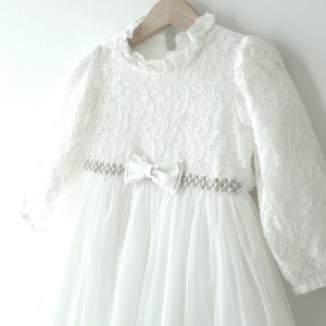 May include: White lace dress with long sleeves and a satin bow at the waist. The dress has a delicate floral pattern and a tulle skirt.