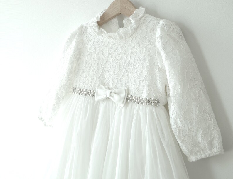 May include: White lace dress with long sleeves and a satin bow at the waist. The dress has a delicate floral pattern and a tulle skirt.